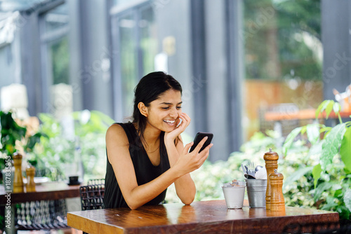 Photography Portrait of smiling, attractive and young Indian woman with her smartphone in a cafe, restaurant or coworking space