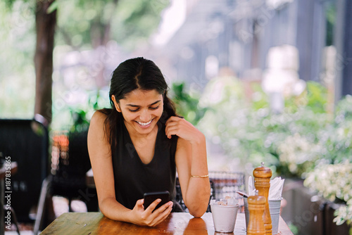 Portrait of smiling, attractive and young Indian woman with her smartphone in a cafe, restaurant or coworking space.  © Danon