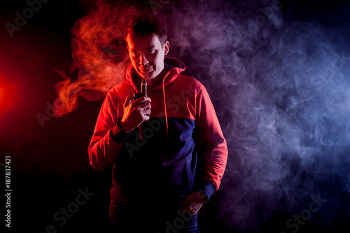 Man in sport hoody vaping an electronic cigarette.Isolated on black background.Around clouds of smoke