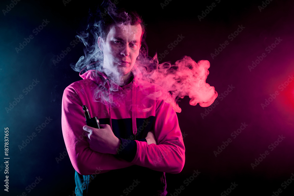 Portrait of a young handsome man holding a  vape and exhaling a large cloud of smoke on a dark background