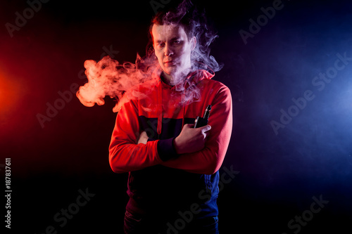 Portrait of a young handsome man holding a vape and exhaling a large cloud of smoke on a dark background
