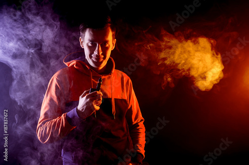 A young stylish man smoker in a hoody is holding an vape in his hand, and around and a big cloud of smoke from vape on a dark isolated background