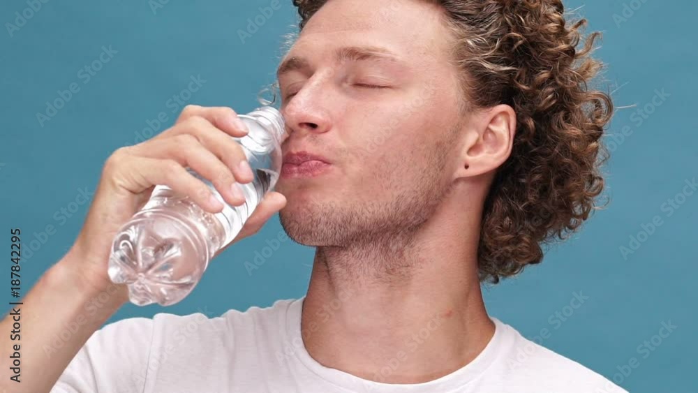 Close up view of Pleased curly man in t-shirt drinking water with closed eyes over blue background Close up view of Pleased curly man in t-shirt drinking water with closed eyes over blue background