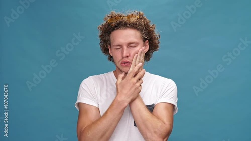 Confused curly man in t-shirt heaving teethache and holding his cheek over blue background
 photo