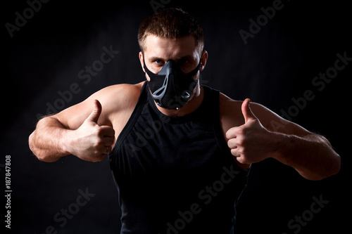 A young athletic man boxer in a sports T-shirt, training black mask shows thumb up on black isolated background