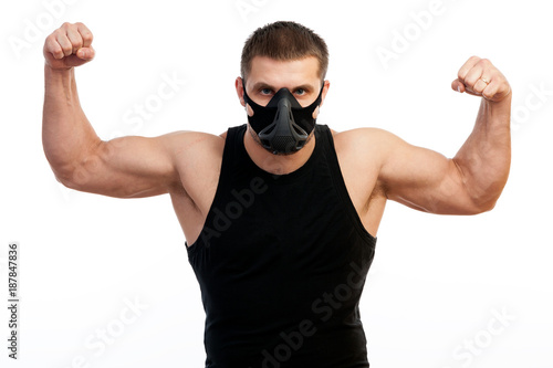 A young athletic man  boxer in a sports T-shirt, training black mask showing his biceps and standing on white isolated background
