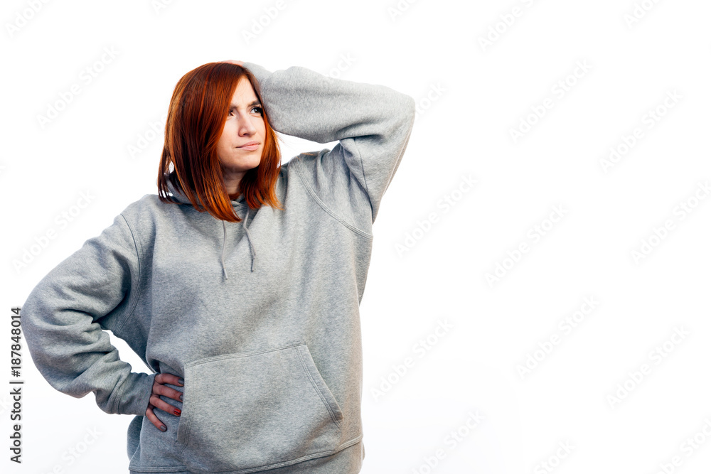 A young red-haired woman in a gray sweatshirt is pensive and holds on to her head with one hand on a white isolated background A young red-haired woman in a gray sweatshirt is pensive and holds on to her head with one hand on a white isolated background