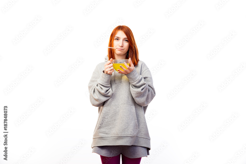 A young red-haired woman in a gray sweatshirt is sick, measures a thermometer temperature and drinks from a large transparent cup of an antiviral agent for treatment on a white isolated background A young red-haired woman in a gray sweatshirt is sick, measures a thermometer temperature and drinks from a large transparent cup of an antiviral agent for treatment on a white isolated background