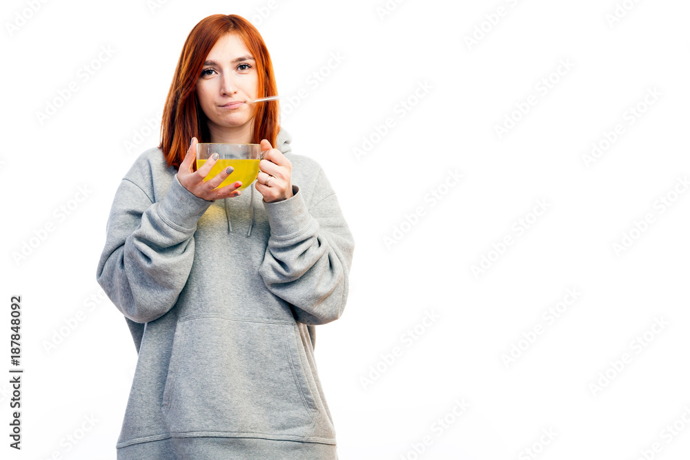 A young red-haired woman in a gray sweatshirt is sick, measures a thermometer temperature and drinks from a large transparent cup of an antiviral agent for treatment on a white isolated background A young red-haired woman in a gray sweatshirt is sick, measures a thermometer temperature and drinks from a large transparent cup of an antiviral agent for treatment on a white isolated background