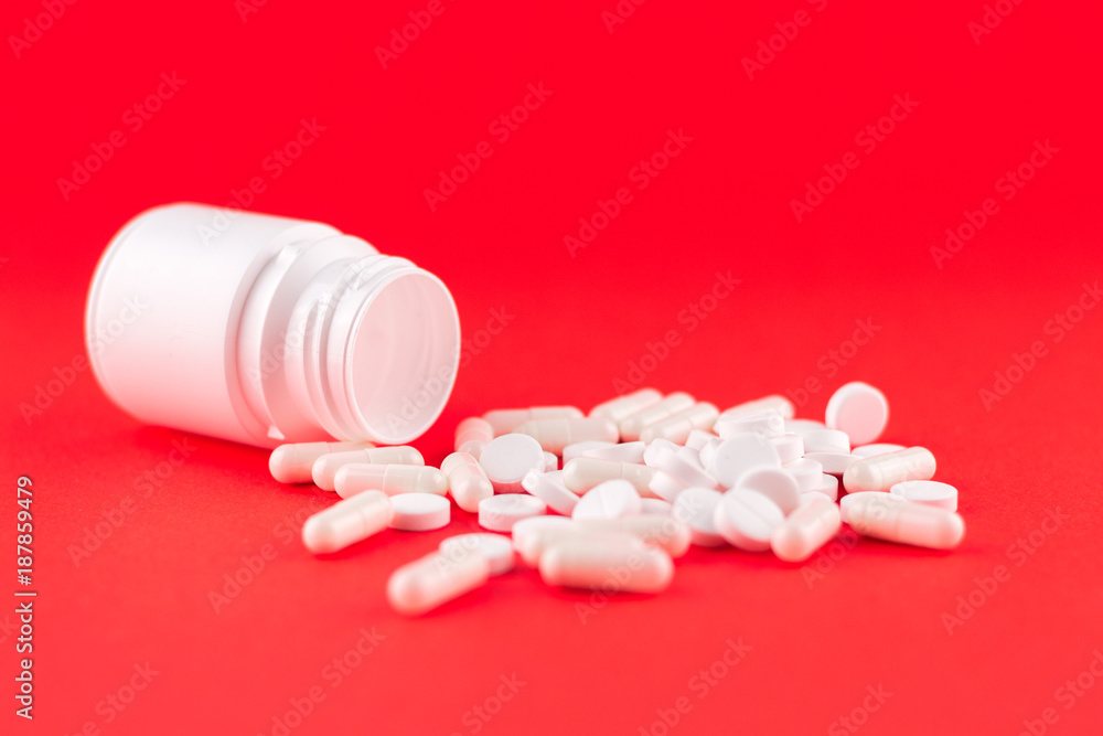 Close up white pill bottle with spilled out pills and capsules on scarlet red background with copy space. Focus on foreground, soft bokeh. Pharmacy drugstore concept Close up white pill bottle with spilled out pills and capsules on scarlet red background with copy space. Focus on foreground, soft bokeh. Pharmacy drugstore concept