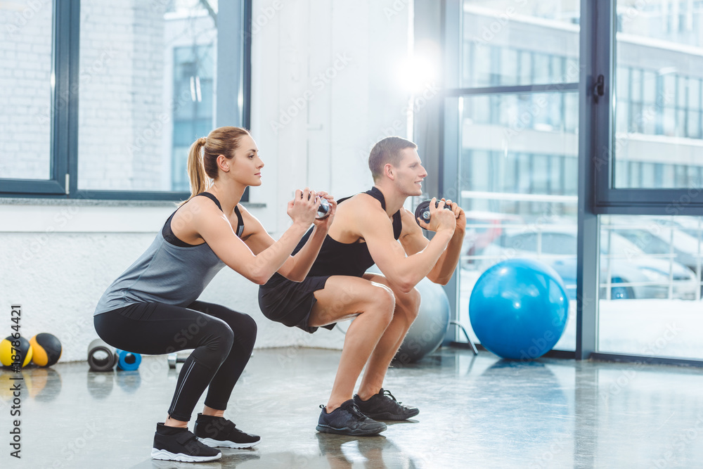 sporty young couple holding dumbbells and doing squat exercise in gym