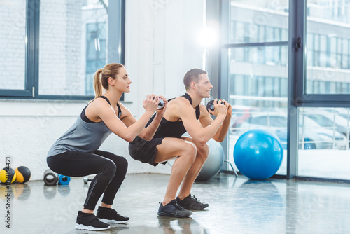 Foto sporty young couple holding dumbbells and doing squat exercise in gym