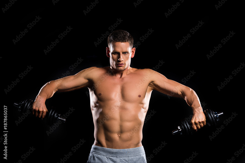 young muscular shirtless sportsman holding dumbbells and looking at camera isolated on black