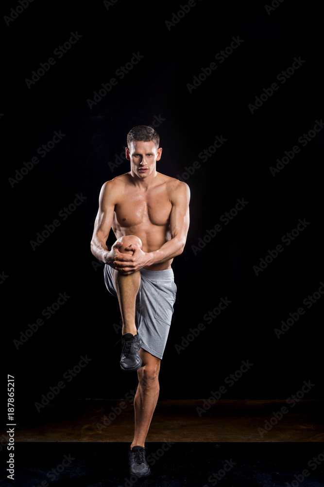 Fototapeta premium muscular young sportsman holding knee in hands and looking at camera on black