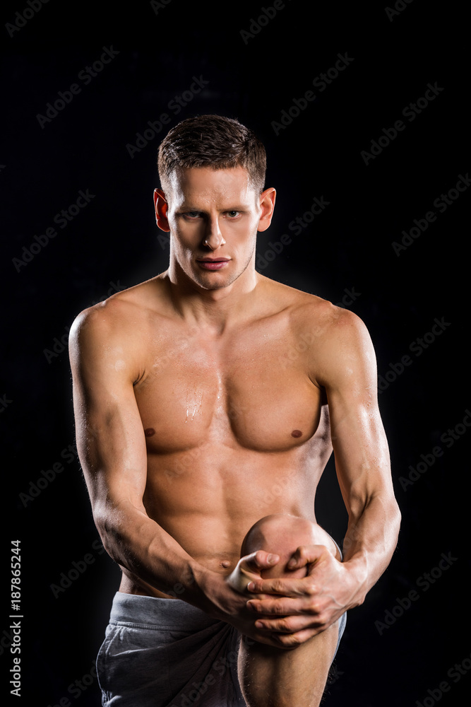 Fototapeta premium muscular young sportsman holding knee in hands and looking at camera isolated on black