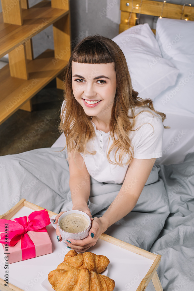overhead view of smiling girl holding cup of coffee in bed