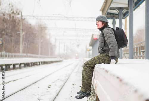 serious young caucasian man with beard sitting at train station in winter in snowy weather