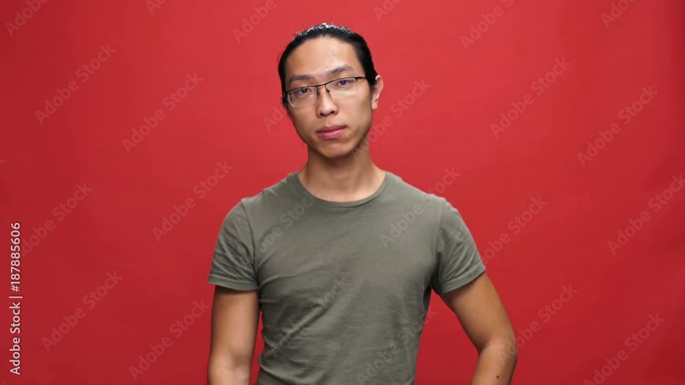 Displeased asian man in t-shirt and eyeglasses disagrees with something and waving his finger over red background Displeased asian man in t-shirt and eyeglasses disagrees with something and waving his finger over red background