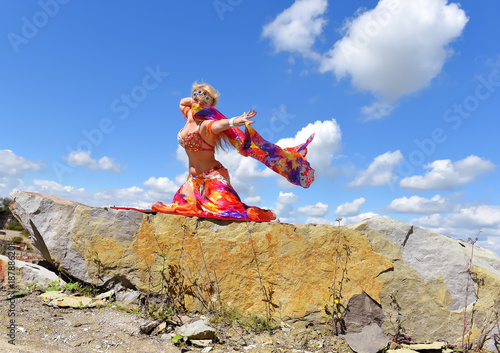 A beautiful blonde oriental  dancer dressed in a multi colored costume,dances  in a large stone quarry. She dances and waves  colorful oriental textiles in the air.