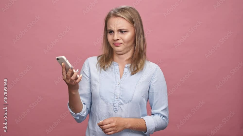 Confused blonde woman in shirt talking by smartphone with someone which screaming on her over pink background Confused blonde woman in shirt talking by smartphone with someone which screaming on her over pink background