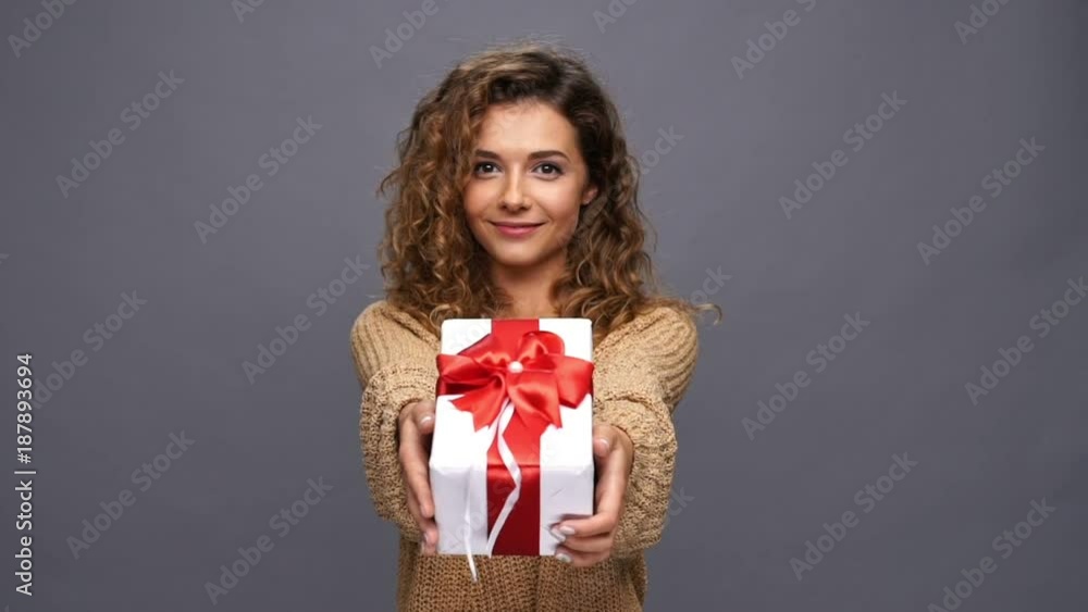 Smiling curly woman in sweater presents a gift box at the camera over purple background Smiling curly woman in sweater presents a gift box at the camera over purple background