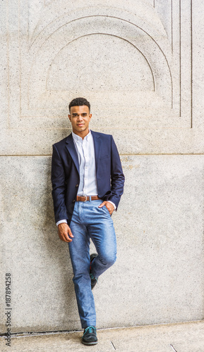 Portrait of African American Man in New York. Wearing blue blazer, white shirt, pants, sneakers, a male college student stands against vintage wall on street on campus, looks at you. Filtered effect..