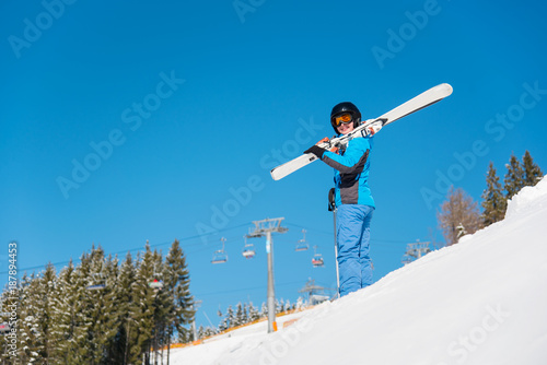 Low angle shot of female skier on the hill in snowy mountains holding her skis on the shoulder. Blue sky, winter forest and ski resort on the background copyspace lifestyle sportswoman weekend concept