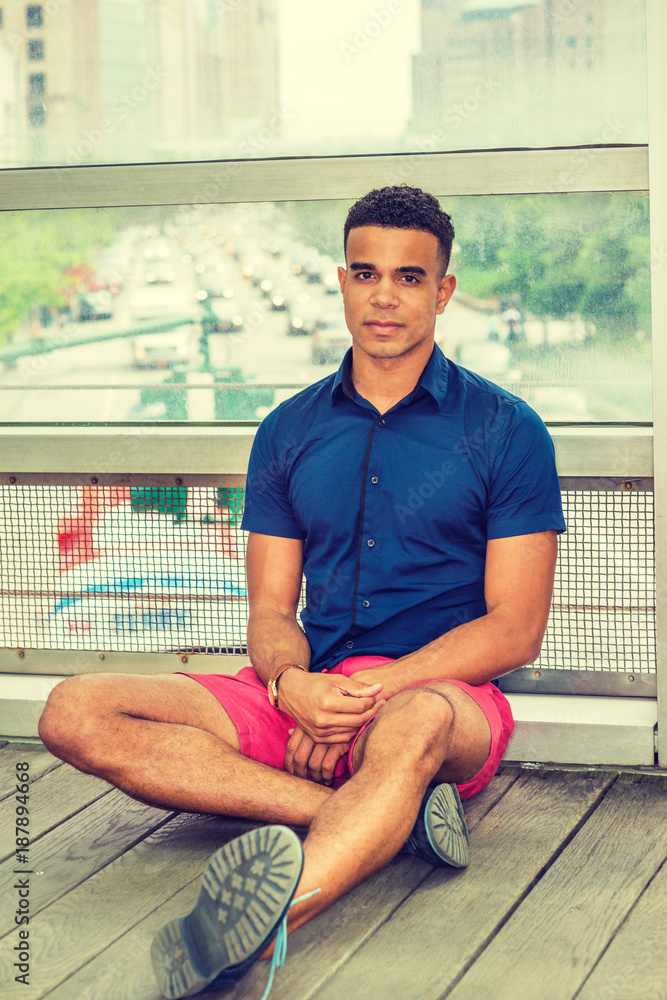 African American man, wearing blue short sleeve shirt, red shorts,  sneakers, sitting cross legged on floor against glass wall on campus,  relaxing, thinking. Street, many cars on background.. Stock-foto | Adobe  Stock