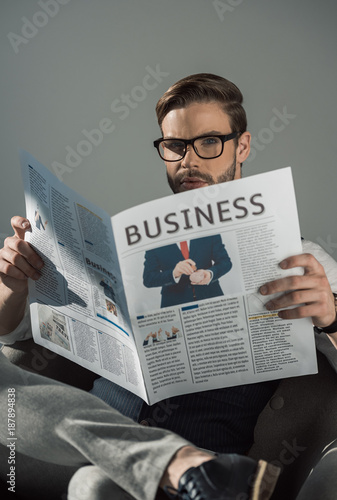 portrait of stylish young businessman in eyeglasses reading newspaper isolated on grey