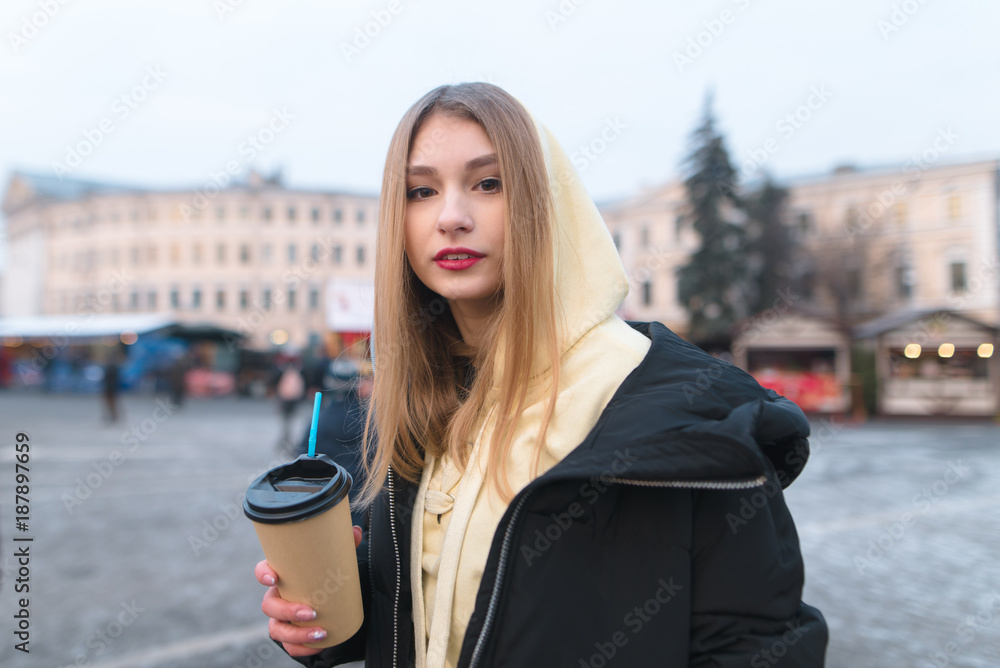 Portrait of a beautiful girl in a jacket with a cup of coffee in her hands against the backdrop of a city landscape. The girl is hot with hot coffee on the street. Portrait of a beautiful girl in a jacket with a cup of coffee in her hands against the backdrop of a city landscape. The girl is hot with hot coffee on the street.