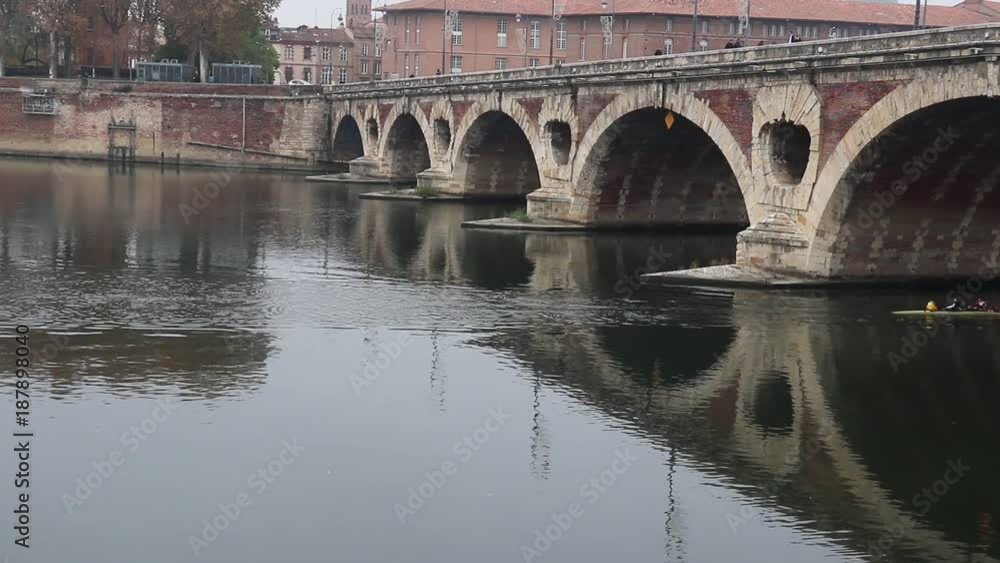 View from the waterfront to the river Garona in the city of Toulouse France. Autumn mood. A team of people in a kayak rowing synchronously and swim under ancient bridge View from the waterfront to the river Garona in the city of Toulouse France. Autumn mood. A team of people in a kayak rowing synchronously and swim under ancient bridge