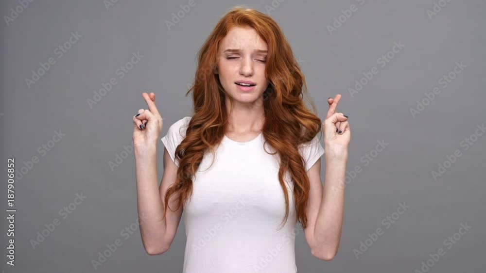 Worried ginger woman in t-shirt praying with crossed fingers over gray background Worried ginger woman in t-shirt praying with crossed fingers over gray background