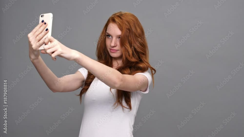 Cheerful ginger woman in t-shirt posing and making selfie on smartphone over gray background Cheerful ginger woman in t-shirt posing and making selfie on smartphone over gray background