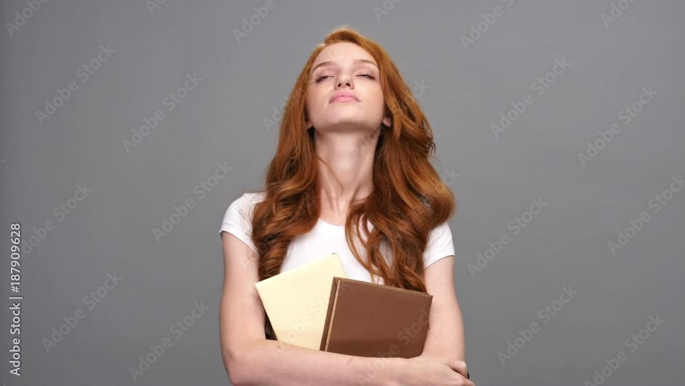 Bored ginger woman in t-shirt holding books and looking around over gray background Bored ginger woman in t-shirt holding books and looking around over gray background