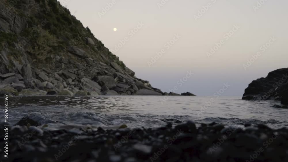 Small waves hitting the shore of a pebble beach at dusk, cliff in the background, moon rising far away. Small waves hitting the shore of a pebble beach at dusk, cliff in the background, moon rising far away.