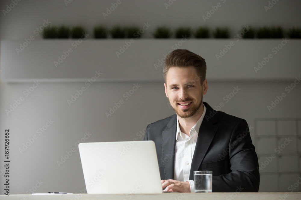 Smiling young businessman in suit working on laptop looking at camera, successful attractive professional executive sitting at office desk using computer, friendly company ceo at workplace, portrait Smiling young businessman in suit working on laptop looking at camera, successful attractive professional executive sitting at office desk using computer, friendly company ceo at workplace, portrait