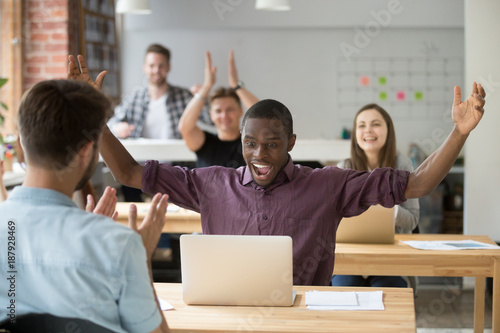 African employee excited about win online raising hands looking at laptop, coworking office team applauding congratulating black colleague with achievement, supporting celebrating success together © fizkes