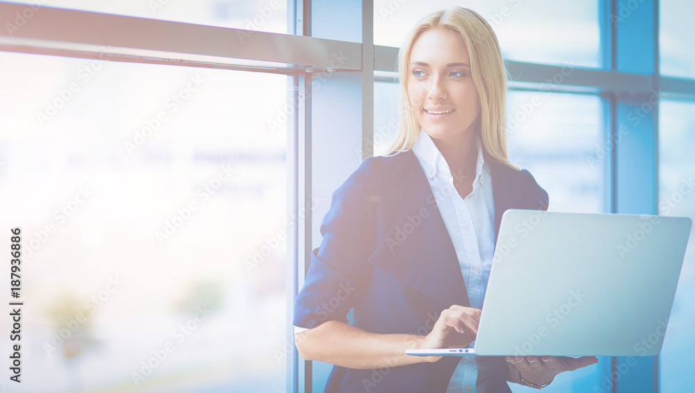 Businesswoman standing against office window holding laptop Businesswoman standing against office window holding laptop