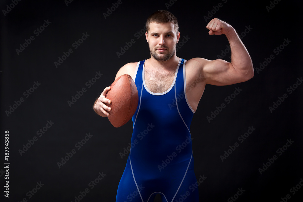 A young dark-haired male wrestler in a blue wrestling tights shows biceps and holds a rugby ball on a black isolated background A young dark-haired male wrestler in a blue wrestling tights shows biceps and holds a rugby ball on a black isolated background