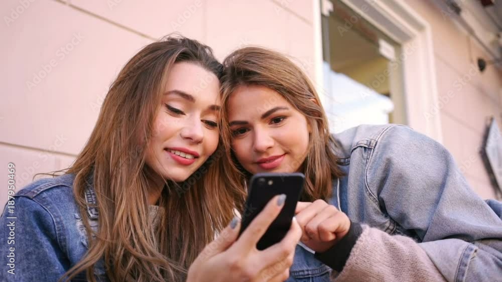 Close up view of Two Pretty smiling women in jeans clothes using smartphone while sitting in city cafe Close up view of Two Pretty smiling women in jeans clothes using smartphone while sitting in city cafe