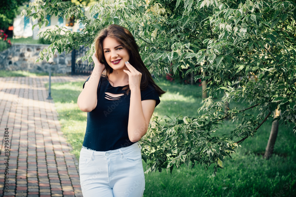 Beautiful model in a black T-shirt and white pants in a park in the middle of green trees.