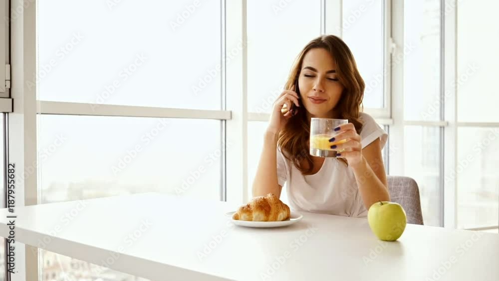 Happy brunette woman sitting by the table with food while talking by smartphone on kitchen at home
Happy brunette woman sitting by the table with food while talking by smartphone on kitchen at home