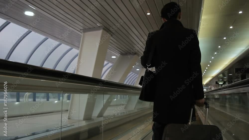 Man in black coat with a suitcase is riding back on airport escalator. Male passenger with luggage slowly moves through a large space to waiting room. Man in black coat with a suitcase is riding back on airport escalator. Male passenger with luggage slowly moves through a large space to waiting room.