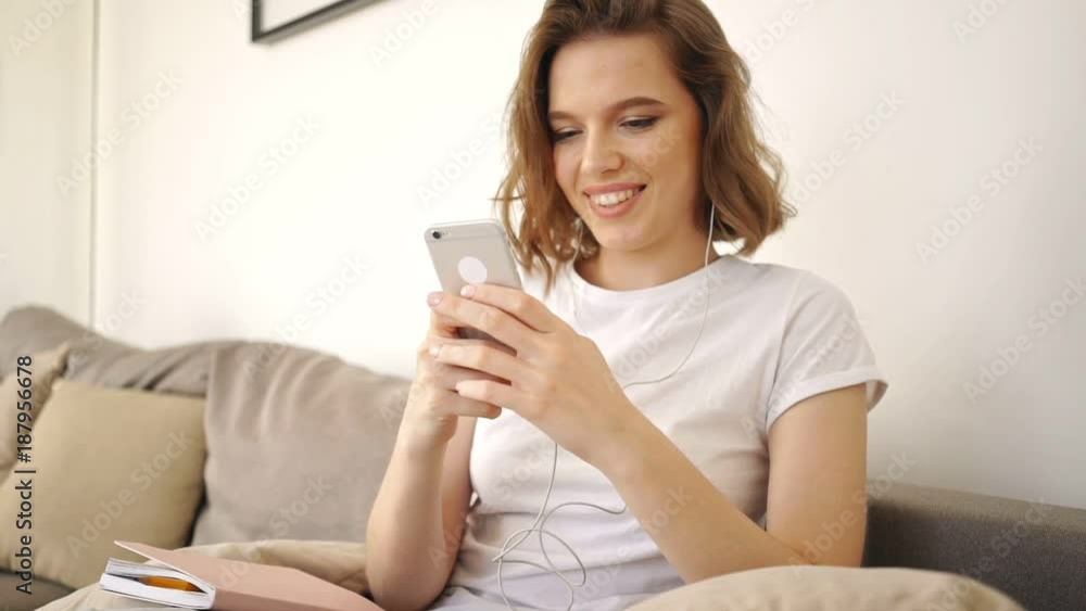 Smiling woman in earphones sitting on sofa and listening music while using smartphone at home Smiling woman in earphones sitting on sofa and listening music while using smartphone at home