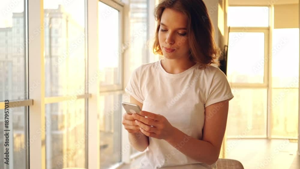 Smiling woman sitting on table and writing message in smartphone on kitchen Smiling woman sitting on table and writing message in smartphone on kitchen