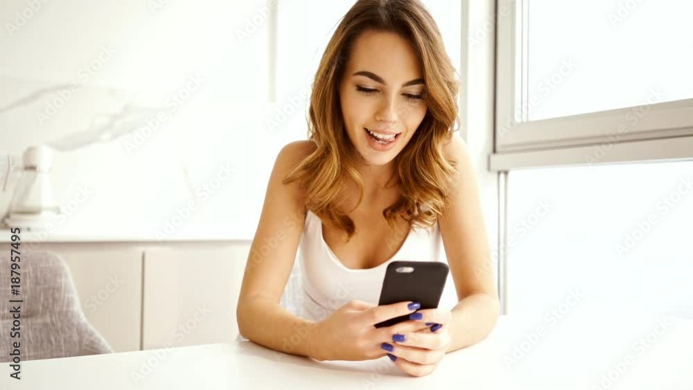 Happy brunette woman sitting by the table while writing message in smartphone at kitchen Happy brunette woman sitting by the table while writing message in smartphone at kitchen