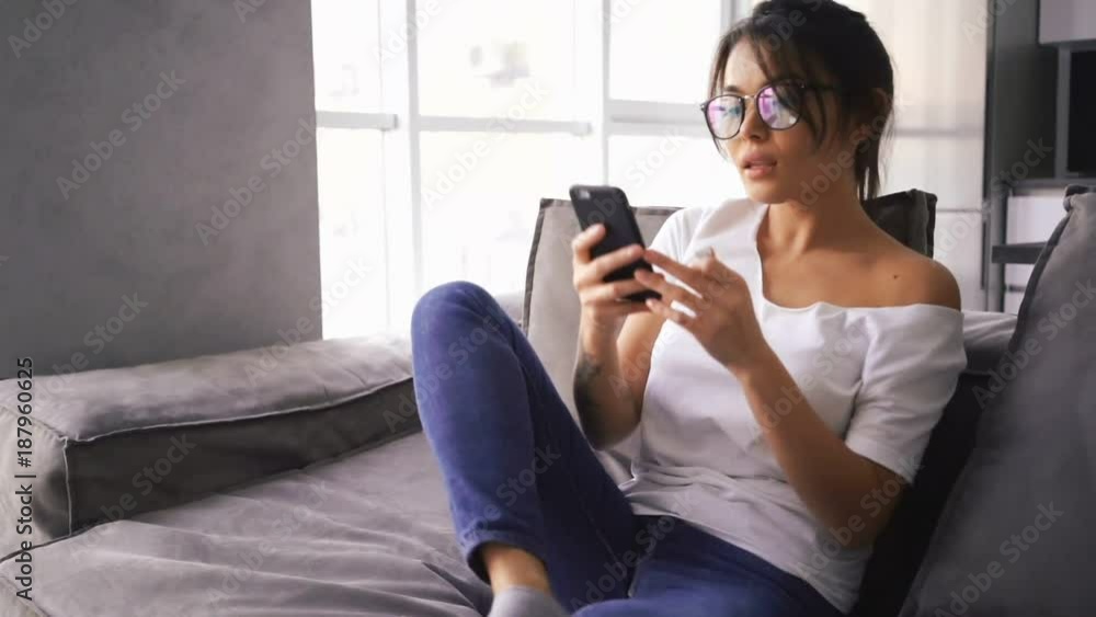 Pretty asian woman in eyeglasses sitting on sofa at home while using smartphone Pretty asian woman in eyeglasses sitting on sofa at home while using smartphone