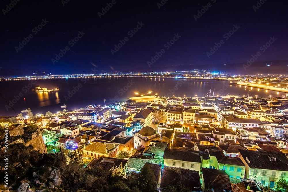 Naklejka premium Illuminated old town of Nafplion in Greece with tiled roofs, small port, bourtzi castle, Palamidi fortress at night.