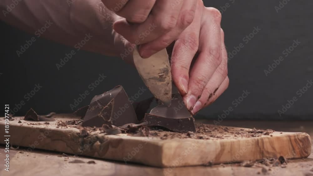 Slow motion closeup young man hands cutting dark chocolate block with parmesan knife on wood board Slow motion closeup young man hands cutting dark chocolate block with parmesan knife on wood board