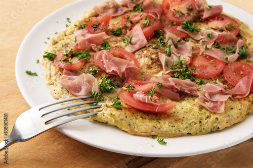 omelette with tomatoes, prosciutto ham and parsley garnish and a fork on a white plate on a rustic wooden table, close up